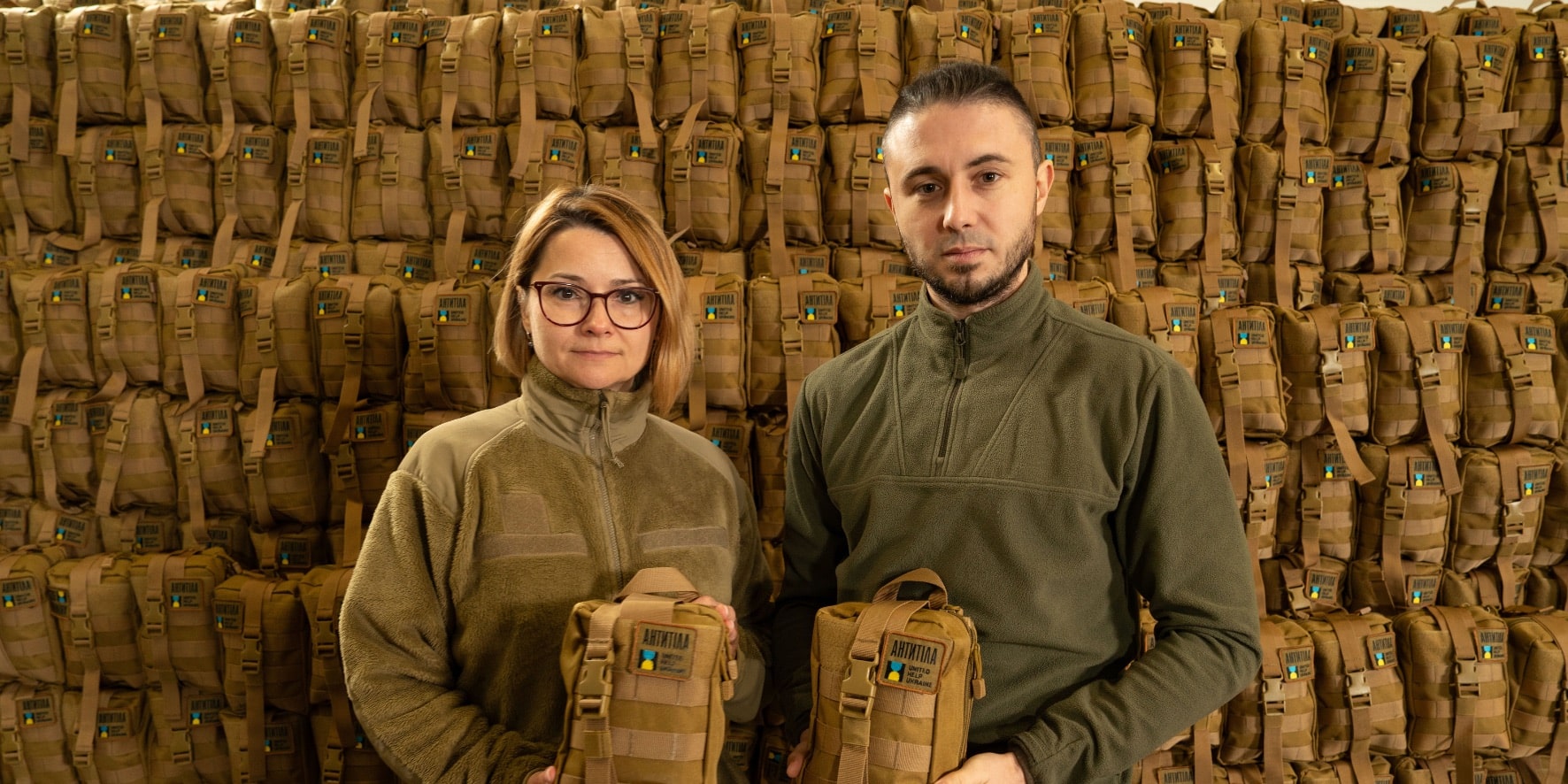 A man and a woman (President of the United Help Ukraine) are holding First Aid Kits in their hands and hundreds of the First Aid Kits on the background.