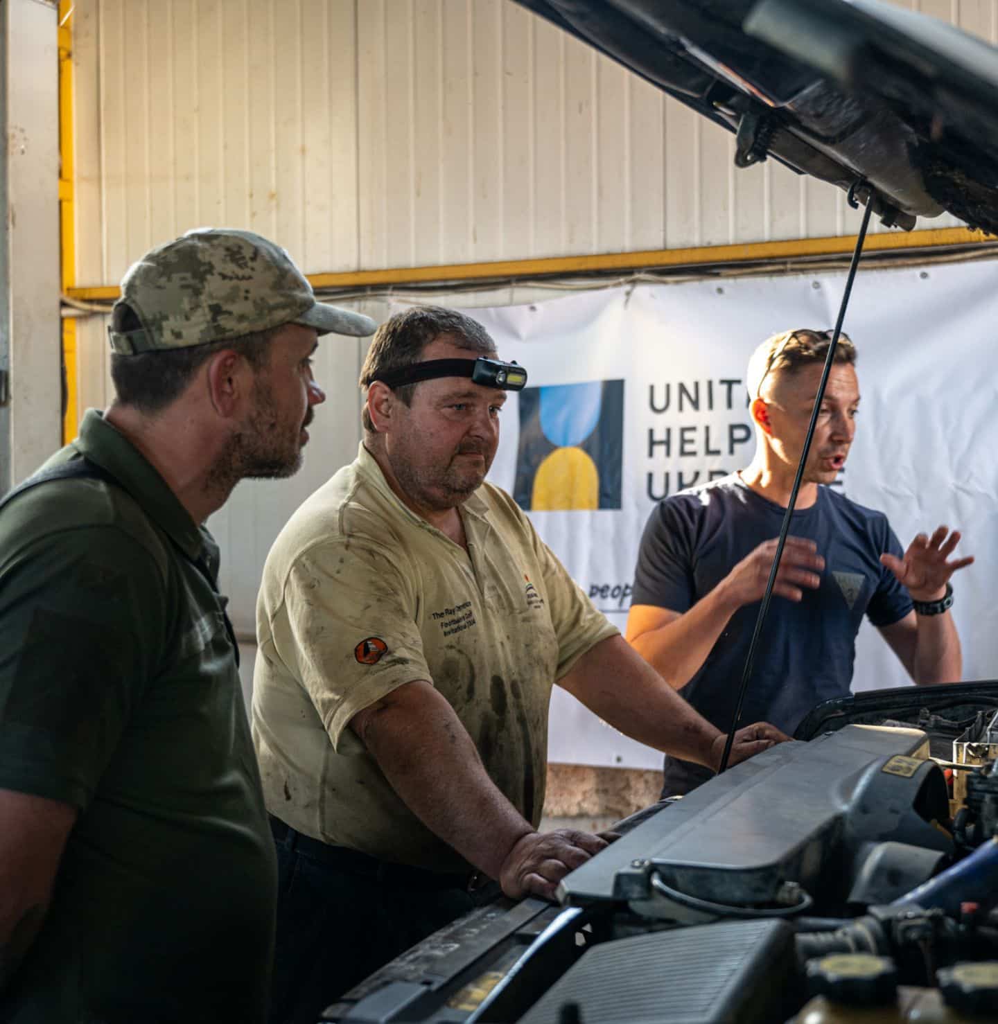 Car mechanics are inspecting a car in their garage to repair it and send it to the front lines. A banner with the United Help Ukraine logo is in the background.
